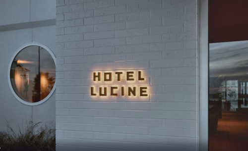 Illuminated sign reading "Hotel Lucine" on a white brick wall, with a round window reflecting the surroundings.